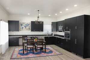 A modern kitchen with black cabinets, stainless steel appliances, and a dining table with four chairs. The table has fruit and flowers on it. A colorful rug is underneath, and a world map decorates the wall. Bright ceiling lights illuminate the room.