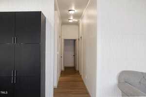 A narrow hallway with light wooden flooring and white walls leads to three doors. The left shows a section of black storage cabinets, and a portion of a gray sofa is visible on the right. Ceiling lights illuminate the space.