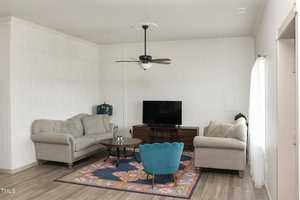 A living room featuring two gray sofas, a wooden coffee table, and a blue chair on a colorful rug. A flat-screen TV is on a wooden stand. A ceiling fan with a light is centered above. The walls are white, and the floor is wood. Natural light enters from a window.