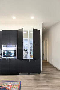 A modern kitchen with black cabinets features an open refrigerator displaying various food items. The cabinet doors on either side of the refrigerator are open. Built-in ovens are installed in the left cabinet. A colorful rug is partially visible on the floor.