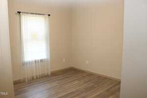 An empty room with light-colored hardwood floors and beige walls. A window on the left with sheer white curtains allows natural light to enter. No furniture or decor is present.