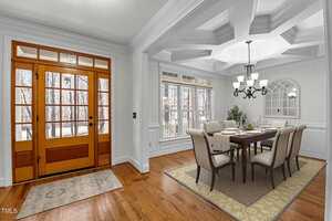 Dining room with a wooden floor and coffered ceiling. A table with six chairs, set with white dishes and green decor, sits under a chandelier. Large windows and a glass-paneled wooden door provide natural light. A patterned rug is near the entrance.