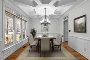 A dining room with a rectangular table and six cushioned chairs. A chandelier hangs above the table. Large windows on the left let in natural light. The walls are light-colored with wainscoting, and there is a framed picture on one wall.