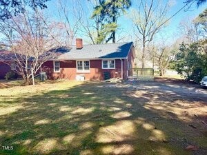 Brick house with dark roof surrounded by trees and a grassy yard. A chimney is visible and there is a wooden deck at the side. A car is parked on the right, partially visible, with a driveway leading to the house.