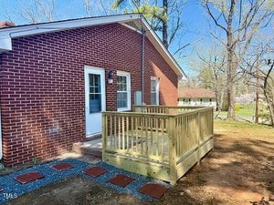 A red brick house with a white door and small wooden deck in the backyard. The ground is bare with scattered leaves and a short path of red bricks. Trees without leaves are visible in the background.