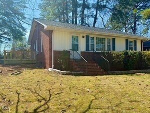 A brick and siding house at 201 W Thorndale Drive in Oxford features a welcoming front porch and steps leading to the entrance. It's surrounded by a grassy yard with shrubs and trees, and a wooden deck is visible on the side. The skies are clear, offering a perfect sunny day backdrop.