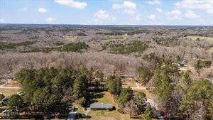 Aerial view of a rural landscape featuring a single-story house surrounded by trees. A mix of forested and open land stretches into the distance under a partly cloudy sky. A few roads and additional buildings are visible.
