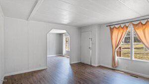 Simple living room with white walls and a light wood floor. A doorway leads to another room with a similar design. Large window with orange curtains, and a door on the right side. Natural light brightens the space.