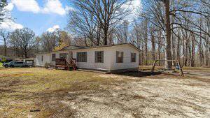 A white, single-story house on 229 County Acres Road in Henderson is surrounded by bare trees, with a wooden porch and swing set. A car rests nearby on the left side, parked on a grassy area beneath a clear sky dotted with clouds.