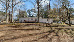 A single-story mobile home is set in a wooded area with tall trees and a brown, leaf-covered yard. A wooden deck extends from the back of the home. A propane tank and a small building are visible nearby.