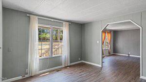 A vacant living room with gray walls and light wood flooring. A large window with white curtains allows natural light in. An arched doorway leads to another empty room with orange curtains. The rooms appear simple and unfurnished.