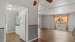 View of an empty room with light wooden floors and white walls. A window with orange drapes is on the right, and an open doorway leads to a kitchen with white cabinets and a white refrigerator on the left. Ceiling fan partially visible above.