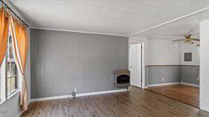 A small room featuring gray walls and wood flooring. An orange curtain covers a window on the left, and a wall heater is mounted. The room opens into another area with a ceiling fan and similar decor. A white door is partially visible.