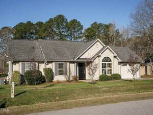 A single-story house with gray siding, a dark gray roof, and a one-car garage. The front yard features a few small trees and shrubs. A small lawn borders the paved street. The sky is clear, and there are trees in the background.