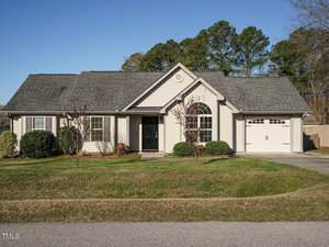 A single-story house at 216 Orkney Road boasts a grey roof and light beige siding. The front lawn, accented by small trees and shrubs, leads to a black front door and a garage with a white door. A large window provides views of the vibrant landscape, where more trees stand in the background.