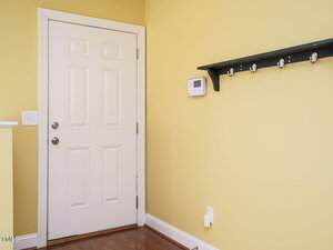 A yellow-walled room features a white door with a silver doorknob and deadbolt. To the right, a black wall-mounted shelf with hooks is visible. A digital thermostat is installed above a white electrical outlet on the wall. Brown wooden floor panels.