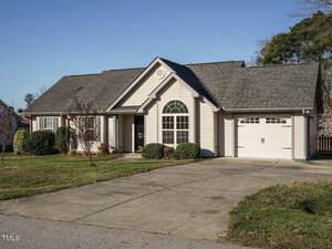 A single-story house with a gray exterior and dark roof. The house features a prominent arched window, a single garage with a white door, and small shrubs in the front. A paved driveway leads to the garage, with a grassy lawn surrounding the property.