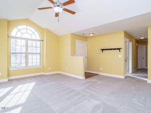 A bright room with yellow walls and carpeted floor. It features a large arched window, a ceiling fan, and recessed lighting. There is a doorway leading to another space and a short wall sectioning off an area with a closed door.