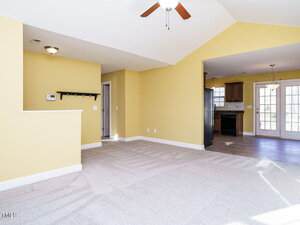 A room with light yellow walls and carpeted floor, featuring a ceiling fan. There is a transition to a tiled kitchen area with dark cabinets and glass-paneled doors leading outside. A white thermostat and coat rack are mounted on one wall.