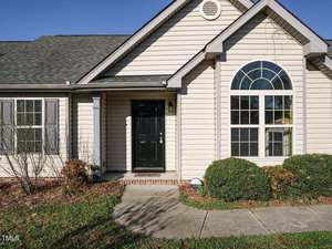Single-story house with light-colored siding, black front door, and arched window with shutters. Low shrubs and a concrete walkway lead to the entrance. The roof is covered with dark shingles, and the sky is clear and blue.