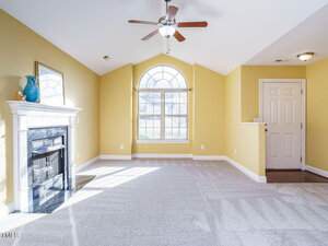A spacious living room with yellow walls and a vaulted ceiling. There is a white-framed fireplace on the left and a large arched window in the center. A ceiling fan is mounted above, and a closed white door is on the right. The floor is carpeted.