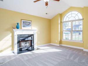A bright living room with yellow walls and a vaulted ceiling. There is a white fireplace with a blue vase and picture frame on the mantel. A large arched window lets in sunlight, illuminating the patterned gray carpet. A ceiling fan is above.