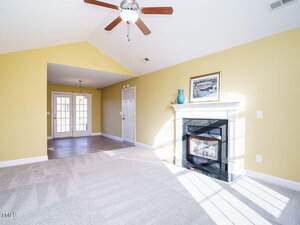 A room with yellow walls and a vaulted ceiling. It has a ceiling fan, a fireplace with a white mantel, and a painting above it. Sunlight enters through glass doors, leading to another room with a hanging light fixture. The floor is carpeted.