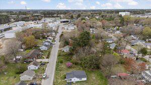 Aerial view of a residential neighborhood with single-story homes, trees, and narrow streets. An industrial area with large buildings and storage tanks is visible in the background. The landscape is lush with greenery and various colored roofs.