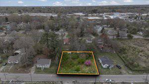 Aerial view of a residential area with a rectangular vacant lot outlined in orange. The lot is overgrown with vegetation and has a pink flowering tree. Surrounding are houses, roads, and trees, with more buildings visible in the background.