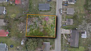 Aerial view of a rectangular plot at 0 Cedar Street with overgrown vegetation, a small building, and a blooming tree. Outlined in orange, the Henderson property is surrounded by houses, a road, and lush greenery.