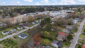 Aerial view of a small residential neighborhood with houses surrounded by trees. There is a large industrial facility in the background. Streets are visible, and several cars are parked along the roads and driveways.
