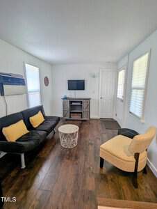 A living room with a dark futon and yellow cushions on the left, a round white coffee table in the center, a patterned yellow armchair on the right, and a TV on a cabinet against the back wall. The room has wooden floors and white walls.