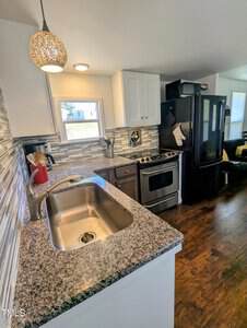 Modern kitchen with granite countertops, stainless steel sink, and tiled backsplash. Features a black refrigerator, white cabinetry, and a stainless steel oven. Hanging pendant light and utensils near the window complete the decor. Dark wooden floor.