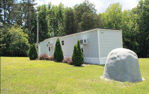 A white mobile home is situated on a grassy lot surrounded by trees. Two small air conditioning units are installed on the side. A large, round gray rock is visible in the foreground. Small, neatly trimmed bushes line the side of the home.