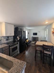 Modern kitchen and living area with granite countertops, stainless steel stove, black refrigerator, and white cabinets. There's a dark wood dining table with chairs, a small couch, and a wall-mounted TV. The room has wooden flooring and a baseboard heater.