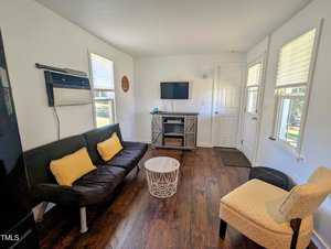 A small living room with a black futon adorned with yellow cushions, an armchair, and a white round table. A flat-screen TV is mounted on the wall above a console. Two windows and a window-mounted air conditioner are visible. Wood flooring throughout.