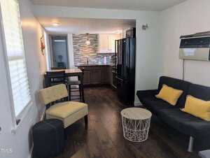 A small living space with a dark floor features a couch with black cushions, a yellow patterned chair, a round white wireframe coffee table, and a dining area with high chairs. The kitchen has a tile backsplash, dark cabinets, and a black refrigerator.