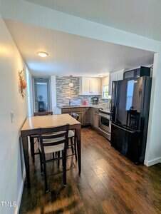 View of a modern kitchen with a center island and two chairs. The kitchen features wooden floors, white cabinets, a black refrigerator, and stainless steel appliances. A decorative piece hangs on the wall, and a window provides natural light.