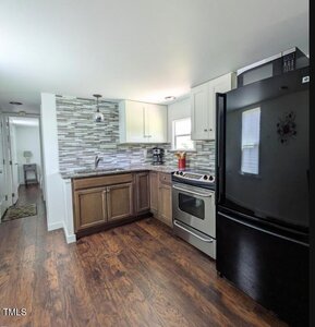 A compact kitchen featuring a black refrigerator, stainless steel stove, and sink. The backsplash is tiled in a gray pattern. Cabinets are white and brown, with wooden flooring. A small window provides natural light, and a hallway is visible in the background.