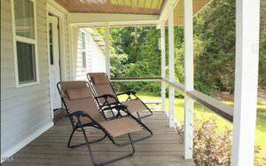 Two brown reclining chairs are positioned on a wooden porch, facing a lush green yard. The porch is attached to a light-colored house, with a partially visible open door. The background includes trees and shrubbery.