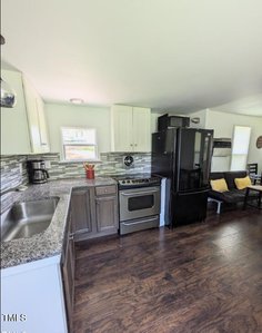 A small kitchen with wood flooring features a stainless steel stove and oven, black refrigerator, and a gray and white tile backsplash. White cabinets line the walls. A window is above the sink, which is set in a speckled granite countertop.