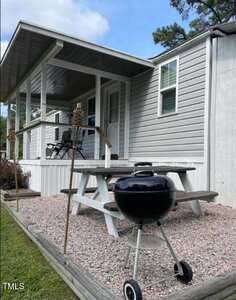 A small wooden porch with a white railing is attached to a gray mobile home. Below, there is a gravel area with a picnic table and a black kettle grill on wheels. Two tiki torches stand on either side of the table. Trees are in the background.