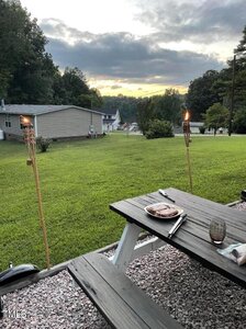 A picnic table is set with a plate and utensils on a grassy lawn. Two tiki torches are lit nearby. In the background, there are houses, trees, and a cloudy sky with the setting sun. Gravel surrounds the picnic area.