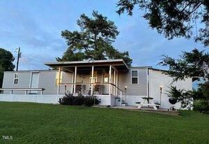 A single-story mobile home with a wooden porch rests on a grassy lawn at 529 Flanagan Road in Henderson. A picnic table and small grill sit on the right side. Pine trees frame the house, and the sky is partly cloudy.