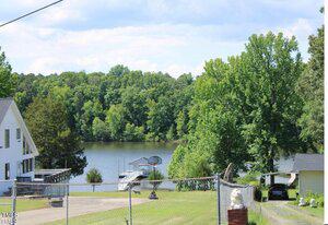 View of a lake surrounded by trees with a small dock and basketball hoop near the shore. A chain-link fence encloses part of the grassy area in the foreground, and a white house and parked car are visible on either side.