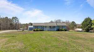 A small, single-story blue house with a covered front porch and a neatly trimmed lawn. The house is surrounded by sparse trees, and a road runs nearby under a clear blue sky.