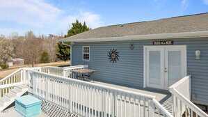 A light blue house with a deck and stairway. The deck has a black metal table and chairs. A sun-shaped decoration is on the wall. Above the door, a sign reads "Bless this Nest." Trees and a shed are visible in the background.