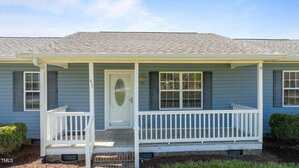 A single-story house with light blue siding and a gray shingled roof. It features a white front door with glass, flanked by two windows with shutters. A white railing frames the small front porch. Trimmed bushes are visible below the porch.