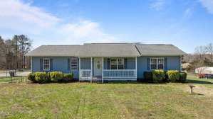 Nestled at 41 Rock Bluff Lane in Kittrell, this charming single-story house boasts blue siding and a gray roof. A cozy front porch with white railings invites you in, overlooking a grassy yard dotted with bushes. The scene is complete with a clear, sunny sky as its backdrop.