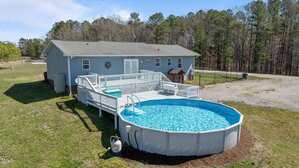 A gray single-story house with a light green roof is shown. In front, a large above-ground pool is surrounded by a white wooden deck. Grass and trees are visible in the background under a clear blue sky.